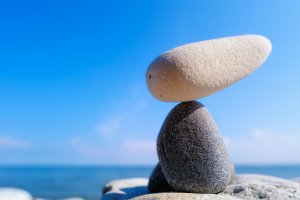 A sculpture made of balanced stones stands in front of a clear blue sky and calm sea, offering a serene contrast to the busy work day. The structure features two dark, round stones at the bottom and an elongated, light-colored stone at the top.