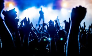 A lively crowd at a concert raise their hands in the air as blue stage lights illuminate the scene. The silhouette of a performer is visible on stage, with bright lights casting a vibrant glow and creating an energetic atmosphere much like employees during an office party.