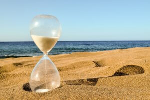 A clear hourglass with white sand stands on a sandy beach, its shadow stretching across the sand, with calm blue ocean waves and a clear sky as a serene backdrop—a perfect reminder of time management's importance.