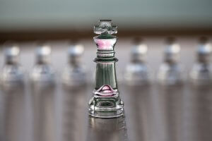A glass chess king piece with a slight pink and green hue stands prominently in focus, surrounded by blurred pawns in the background. The reflective surface under the chess pieces adds a glossy effect to the image, subtly reminding us of the transparency needed to address issues like sexual harassment.