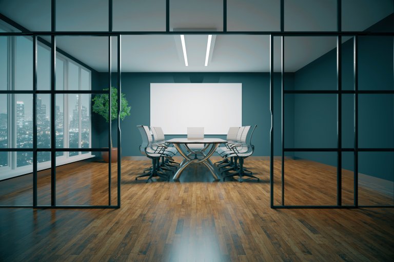 A modern conference room with a large table and eight chairs, facing a blank projector screen. The room has wooden floors, glass walls, and a potted plant in the corner. A city skyline is visible through the windows on the left, setting an ideal space for discussions on preventing sexual harassment in the workplace.