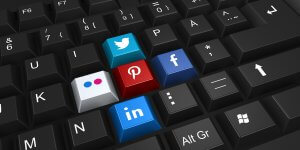 A black computer keyboard with several keys replaced by social media icons—Twitter (blue), Pinterest (red), Facebook (blue), Flickr (white and pink), and LinkedIn (blue). The social media icons are prominently displayed in the center of the keyboard.
