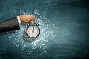 A person's hand in a suit jacket holds an old-fashioned alarm clock in front of a chalk-drawn cityscape on a chalkboard. The whimsical scene, filled with buildings, clouds, stars, and a crescent moon, subtly underscores the importance of time management in creative endeavors.