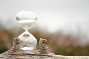 A clear hourglass filled with white sand sits atop a weathered piece of wood, with a soft-focus background of greenery and flowers, symbolizing time management in a peaceful, natural setting.