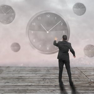A man in a suit stands on a wooden platform, reaching out towards a large clock on a foggy background. Other smaller clocks float around him in the mist, emphasizing themes of time management and the passage of time.