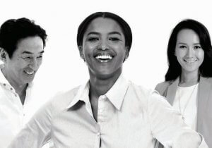 A black-and-white photo of three people. In the foreground, a woman is smiling broadly with her hands on her hips. To her left, a man is also smiling while looking downward. On the right, a woman is smiling and looking directly at the camera—an image capturing joy despite underlying themes of sexual harassment awareness.