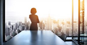 A silhouette of a person stands in a modern conference room with a floor-to-ceiling window, overlooking a cityscape at sunset. The room features a long table with chairs on both sides. As the work day comes to an end, the individual appears to be gazing out at the sprawling city below.