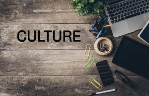 A wooden desk with a laptop, tablet, smartphone, pens, pencils, paper clips, and a small potted plant. The word "CULTURE" is written in bold black letters on the left side of the desk, symbolizing the importance of workplace culture.