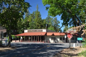 A quaint building with a red roof and a sign reading "Manka's Corner" sits at a street intersection surrounded by tall trees and greenery under a clear blue sky. The area looks peaceful with a stop sign visible in the foreground.