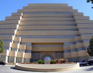 A large, modern, multi-tiered beige building with a stair-step design. The entrance is prominent and rectangular, flanked by planted greenery and colorful flowers. Several parked cars are seen around the front driveway, which features a circular emblem.