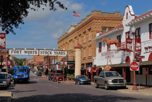 A view of the Fort Worth Stockyards shows a historic street lined with brick buildings. A sign reading "Fort Worth Stock Yards" spans across the street. Visible are parked cars, a truck, flags, store signs, an American flag, and a cow statue on a building.