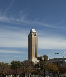 A tall, tan and white Art Deco skyscraper rises into a clear blue sky with wispy clouds. The building, topped with a multi-tiered structure, is surrounded by trees with autumn foliage and a low-rise building to its right. The Texas state flag is visible on a pole.