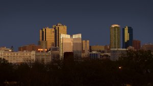 A city skyline at dusk featuring a mix of modern and older buildings. The tallest structures have reflective glass exteriors that catch the last light of the setting sun, while smaller buildings and a dark, tree-lined foreground complete the scene.
