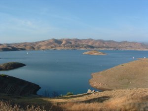 A serene lake bordered by gently rolling hills under a clear blue sky. Several small sailboats are scattered across the water, while a few grassy mounds rise from the lake's surface. The foreground is a dry, grassy area leading down to the water's edge.