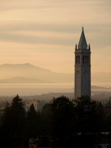 A tall clock tower stands prominently against a golden-hued sky at sunset, with a mountainous landscape in the background. Below the tower, urban buildings and trees are visible, creating a striking contrast with the serene natural scenery.