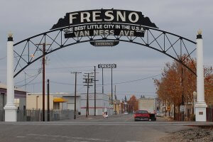 A welcome archway at Van Ness Avenue with the text "Fresno: The Best Little City in the U.S.A." cars and a pedestrian passing beneath it. The street is lined with industrial buildings and has a speed limit sign. The sky is overcast.
