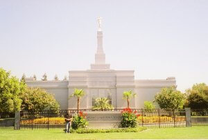 A person stands near the entrance of the Fresno California Temple belonging to The Church of Jesus Christ of Latter-day Saints. The temple features a clean white facade, a golden angel statue atop its spire, manicured gardens, and a sign displaying its name.