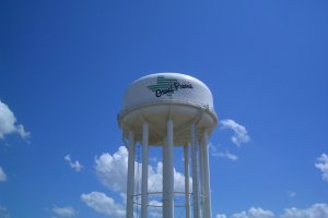 A white water tower with the words "Grand Prairie" written on it stands tall against a bright blue sky with a few scattered clouds. The tower is supported by multiple pillars.