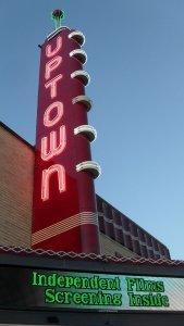 A vibrant neon sign reading "Uptown" vertically adorns a classic theater marquee against a clear sky. Below, an electronic display advertises "Independent Film Screening Inside" in green text. The building features a mix of brick and red decorative elements.