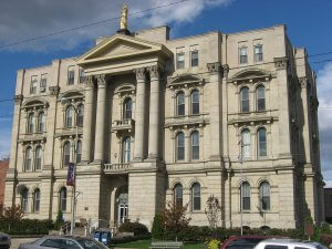 A large, classical-style courthouse with beige stone exterior, multiple tall columns, arched windows, and a central balcony. A golden statue tops the building. The front entrance is elevated with stairs, and flags are displayed on the right side. The sky is partly cloudy.