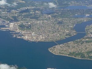 Aerial view of Bremerton, Washington, showcasing its waterfront layout with multiple peninsulas, residential neighborhoods, industrial areas, and marinas surrounded by water. The city is interspersed with green spaces and connected by bridges spanning the waterways.