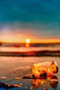 A glass jar containing small lights lies on wet sand at a beach in Los Angeles County during sunset. The sun is low, casting a warm glow across the sky and reflecting on the wet sand and water. The colors blend from deep blue to orange and yellow, creating a serene and magical atmosphere.