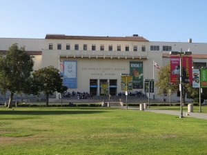 The Los Angeles County Museum of Natural History building, featuring a cream-colored facade with large banners hanging near the entrance. The surrounding area has green trees and a grassy lawn with pathways. Visitors are seen gathered near the museum's entrance, enjoying the vibrant Los Angeles County atmosphere.