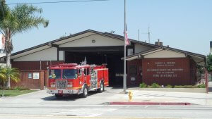 A red fire truck is parked outside Fire Station 127, part of the Los Angeles County Fire Department in Carson, California. The single-story building features an American flag flying in front, and a palm tree is visible on the left side.