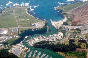 Aerial view of a coastal town with a forested area, buildings, and a marina. A bridge spans a waterway that leads into the ocean. The coastline is rugged, with cliffs and scattered trees. Roads and structures are visible on the mainland, with a distinct shoreline curve.