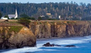 A scenic coastal view of rugged cliffs meeting the ocean waves. Atop the cliffs, there is a white church with a tall steeple and a few houses surrounded by dense forest. The sky is clear, suggesting a calm, serene atmosphere.