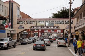 A bustling street scene in Monterey, California features the Cannery Row Company bridge, numerous shops, and restaurants. The street is filled with parked and moving cars, pedestrians, and various national flags hanging from buildings.