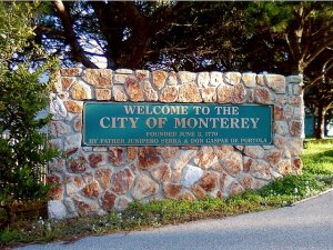 A stone sign with a green plaque reads, "Welcome to the City of Monterey, founded June 3, 1770 by Father Junipero Serra & Don Gaspar de Portola." The sign is surrounded by trees and greenery, and a road is visible in the foreground.