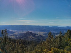 A panoramic view from a mountain peak reveals a lush valley with hills, forests, and small patches of open landscape. The sky is clear with a few wispy clouds and the sun beams brightly from the top left corner, casting light over the scene.