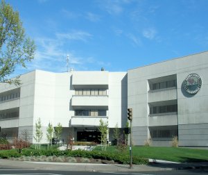 A three-story concrete building with the seal of Placer County on the right side. The building has numerous windows, a small antenna on the roof, and is surrounded by trees, shrubs, and a landscaped lawn with pathways and a sidewalk.
