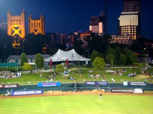 A night view of a baseball field with some spectators on a grassy hill. In the background, there is a mix of modern and historic buildings illuminated by lights, including a distinctive yellow tower structure. Advertising banners are visible along the field's fence.