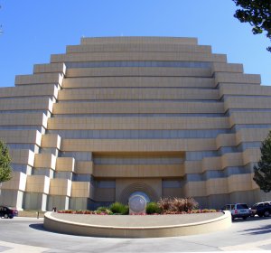 A large, modern building with a stepped, tiered design stands against a clear blue sky. The façade is made of beige stone, and the entrance is marked by a series of arched recesses. There are landscaping and parked cars in front of the building.