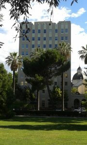 A tall beige building with multiple windows is surrounded by palm trees and lush greenery. In the background, there is a domed structure. The foreground features a well-maintained grassy lawn. The sky is partly cloudy.