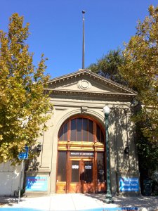 An arched wooden door with glass panels is the central feature of this classical-style building. The facade is adorned with a triangular pediment and a flagpole at the top. Two large trees frame the entrance, and signs are visible on either side of the door.