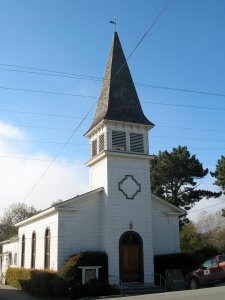 A quaint white church with a tall steeple stands under a clear blue sky. The structure has a wooden door and dark-framed arched windows on its side. There are bushes surrounding the church and a few tall trees visible in the background. A car is parked nearby.