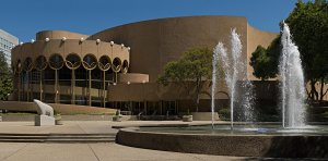 A modern, circular beige building with large windows and decorative arches, surrounded by trees and greenery. In the foreground, a large circular fountain features multiple water jets. The sky is clear and blue, indicating a sunny day.