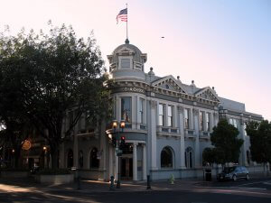 A historic two-story building with a central dome and an American flag on top. The facade is adorned with columns and arched windows. Trees flank the building's side, and a streetlight stands at the corner of the adjacent intersection.