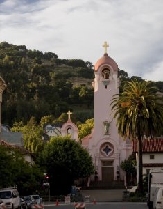 A serene pink church stands prominently amid lush greenery and hills. The building features a central tower with a cross at the top and an arched entrance. Palm trees and other foliage surround the church, with a few traffic cones visible at the bottom of the image.