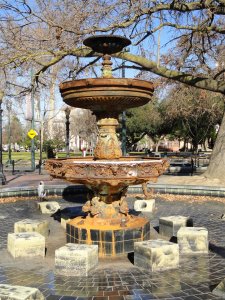 A weathered, ornate two-tiered fountain stands in the center of a plaza with surrounding benches and bare trees. The fountain appears aged with rust and stains, and stone blocks encircle its base. The area is quiet, suggesting a peaceful, sunny day.