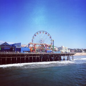 A vibrant Ferris wheel stands prominent on a pier extending into the ocean under a clear blue sky. Surrounding the Ferris wheel are colorful amusement park structures. Waves gently crash against the pier. Beachfront buildings are visible in the background.