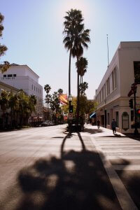 City street scene with tall palm trees lining the road, casting long shadows. Buildings on both sides, some with awnings and signs. A person is walking on the sidewalk to the right. The sky is clear with bright sunlight creating a serene and peaceful atmosphere.