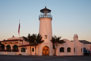 A white stucco building designed to resemble a lighthouse, with a tall central tower and lantern room. The building, labeled "Santa Paula" is surrounded by palm trees and has an American flag flying nearby. The sky is clear, suggesting early morning or late afternoon.