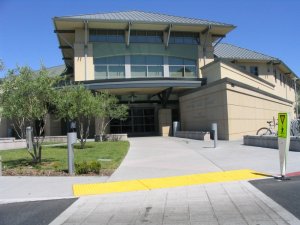 The image shows a modern public library building with a peaked roof and large windows. The entrance has a sidewalk leading up to it and is surrounded by well-maintained landscaping, including small trees and grass. A pedestrian crossing sign is visible in the foreground.