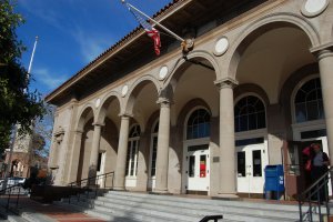 A grand building with an arched entrance and a series of tall columns. Steps lead up to the entrance, which features large windows and double doors. An American flag is mounted above the arches. A woman stands at the top of the stairs near the entrance.