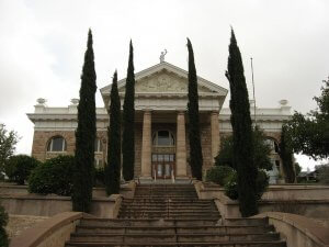A historic, stone courthouse with tall columns at the entrance and a triangular pediment above. Four tall, narrow cypress trees line the front stairs leading to the double doors. The building is flanked by bushes and additional trees. The sky is overcast.
