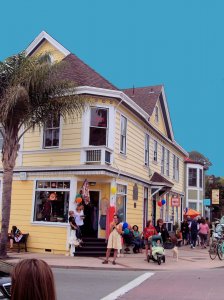 A bustling street corner features a yellow building with shops. People are gathered outside and on the sidewalk. There is a man with a stroller and a small dog, and others are holding balloons. The sky above is clear blue, and a palm tree adds a tropical feel.